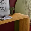 A close-up of a rattan console table with a book and a lamp on it, set against a backdrop of dark red and light green walls. Produktbild-Vorschau