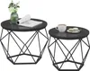 Two round coffee tables with geometric black metal bases are shown against a white background. The larger table on the left has a green vase with light green flowers and a rolled-up magazine on it. The smaller table on the right has a white mug of coffee a Podgląd zdjęcia produktu