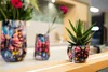 Three decorative pots with a butterfly design are displayed on a wooden surface, with a succulent plant in the smallest pot and flowers in the largest. Produktbild-Vorschau