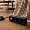 A black power strip with a red illuminated switch is lying on a wooden floor next to a beige knitted blanket. Podgląd zdjęcia produktu