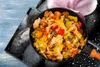 A close-up of a dish of food, possibly a stir-fry, in a black pan, set against a backdrop of black stones with water droplets and a pink flower petal. Náhled obrázku produktu