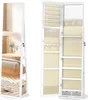 A white jewelry cabinet with a full-length mirror is shown open, revealing its interior storage compartments and shelves. The cabinet is placed in a bedroom setting, with a bed and nightstand visible in the reflection of the mirror. Náhled obrázku produktu
