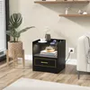 A black nightstand with a gold drawer pull is shown in a bedroom setting, next to a couch and a plant. Náhľad obrázku produktu