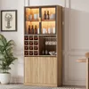 A tall, multi-compartment cabinet with glass doors and interior lighting, designed for storing wine and glassware, placed in a room setting with a plant and a framed picture. Náhled obrázku produktu
