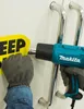 A person wearing gloves is using a heat gun to remove a yellow sticker with black text from a white surface near two silver door handles. Podgląd zdjęcia produktu