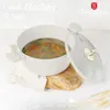 A white pot with a glass lid, filled with soup, is placed on a wooden board and a white cloth. The background is a light gray textured surface. Text and logos are present in the image. Produktbild-Vorschau