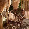A macrame hanging chair with cushions sits on a wooden deck, surrounded by pampas grass and flowers. Produktbild-Vorschau