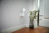 A white LED plug-in night light with a flashlight function is mounted in an electrical outlet on a light gray wall. Next to it, a glass vase with green foliage stands on a wooden floor. Produktbild-Vorschau