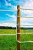 A metal gate attached to a wooden post in a grassy field with a blue sky and clouds. Produktbild-Vorschau
