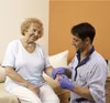 A caregiver is examining an elderly woman's hand while she smiles. Produktbild-Vorschau