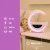 A bedside table with a modern lamp that functions as a clock, Bluetooth speaker, and wireless charger. A doll is visible in the background. Náhled obrázku produktu
