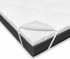 A white mattress topper is shown on top of a dark gray mattress. The topper has a diamond-stitched pattern and elastic straps on the corners to secure it to the mattress. Produktbild-Vorschau