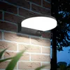 A black LED wall lamp with a white light panel is mounted on a brick wall at night. Some green leaves are in the foreground. Podgląd zdjęcia produktu