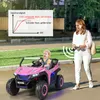 A woman is remotely controlling a pink ride-on toy car with a child inside, outdoors on a paved area next to a grassy verge. Produktbild-Vorschau