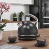 A black retro-style electric kettle is shown on a wooden countertop in a kitchen setting, with two cups of tea and a plant in the background. Produktbild-Vorschau