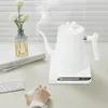 A white electric kettle is shown on a white base, with steam rising from its spout. A vase with flowers and a book are on the left. A hand is reaching for a mouse in the foreground. Náhled obrázku produktu