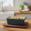 A casserole dish filled with food sits on a wooden cutting board on a kitchen counter. Produktbild-Vorschau