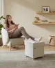 A woman is sitting in a chair reading a book, with her feet resting on an ottoman. The scene is set in a living room with shelves on the wall. Produktbild-Vorschau