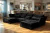 A spacious living room with a modern black sectional sofa in the foreground and a kitchen area in the background.
