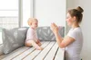 A mother and baby are sitting on a wooden bench by a window, with pillows behind them. The mother is clapping her hands and smiling at the baby, who is looking at her. Produktbild-Vorschau
