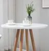 A white round table with wooden legs, set with two coffee cups and saucers, and a vase with greenery. Podgląd zdjęcia produktu