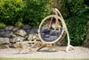 A wooden hanging chair with a gray cushion sits on a gravel patio next to a small wooden table with drinks, in a garden setting with a stone wall and lush greenery. Produktbild-Vorschau