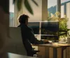 A person working at a desk with dual monitors in a well-lit office with a view of nature. Produktbild-Vorschau