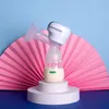 A white electronic breast pump with a clear bottle containing beige liquid is displayed on a white circular stand. Behind it is a pink folded paper fan, and the background is split between light blue and dark blue. Podgląd zdjęcia produktu