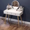 A white vanity table with a round mirror and wooden legs, set against a dark blue wall and wooden floor. It is decorated with makeup and a small vase of flowers. Náhľad obrázku produktu