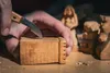 A close-up shot of a person's hands carving a small wooden block with a knife, with other carved wooden figures in the background. Produktbild-Vorschau