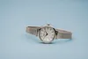 A silver wristwatch with a white face is displayed on a light blue background. Produktbild-Vorschau