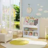 A child's bedroom with a white bookshelf filled with books and toys, a white armchair, and a large window overlooking a garden and lake. Produktbild-Vorschau