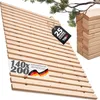 A wooden slatted base, likely a bed slatted frame, is displayed diagonally against a white background. A branch with green leaves is visible in the upper right corner. A stack of similar wooden slats is also in the background. Produktbild-Vorschau