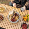 A person is holding a cup and sitting at a wooden table with a portable grill and various food items. Anteprima dell’immagine prodotto