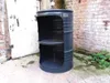 A modified oil barrel used as a shelf unit, placed outdoors next to a brick wall and a white door. Náhled obrázku produktu