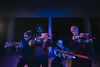 Four people holding toy guns in a dimly lit, possibly urban, environment with blue and purple lighting. Voir les images du produit