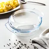 A clear glass casserole dish with a lid sits on a light gray surface, next to a plate of rolled pasta and a napkin. Produktbild-Vorschau