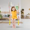 A young girl is swinging indoors on a wooden swing with yellow handles and red beads on the ropes. The background shows a living room with furniture and decor. Produktbild-Vorschau