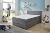 A bedroom scene with a boxspring bed, a coat rack, a suitcase, and decorative items on the wall. Produktbild-Vorschau