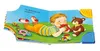 A children's book open to a page showing a baby playing with toys on a grassy surface. Produktbild-Vorschau