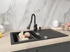 A modern kitchen sink with a black granite basin, a flexible black faucet, and a selection of fruits in a drying rack. Produktbild-Vorschau