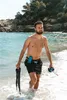 A man walks out of the ocean on a sunny day, holding snorkeling gear. Podgląd zdjęcia produktu