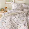 A close-up view of a bed with floral patterned bedding and a bedside table with a lamp and plant. Produktbild-Vorschau
