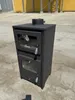 A black wood-burning stove with a glass door and a chimney pipe on top, placed outdoors on a concrete surface with yellow structures in the background. Produktbild-Vorschau