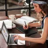 A young woman is using a graphics tablet and stylus connected to a laptop, with books and a mug on the table. Produktbild-Vorschau