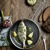 A cooked fish in a pan with lemon slices and herbs, served on a wooden table with bread and an artichoke. Náhľad obrázku produktu