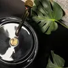 A close-up overhead view of a black basin with a gold faucet and a large green leaf. Produktbild-Vorschau