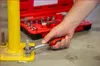 A person is using a ratchet to tighten a bolt on a yellow metal post. A red toolbox with tools is in the background. Náhled obrázku produktu