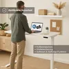 A man is standing at a white height-adjustable desk, using a laptop. The desk has features highlighted with text overlays. Produktbild-Vorschau