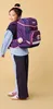 A young girl wearing a backpack and school uniform, posing against a plain background. Produktbild-Vorschau